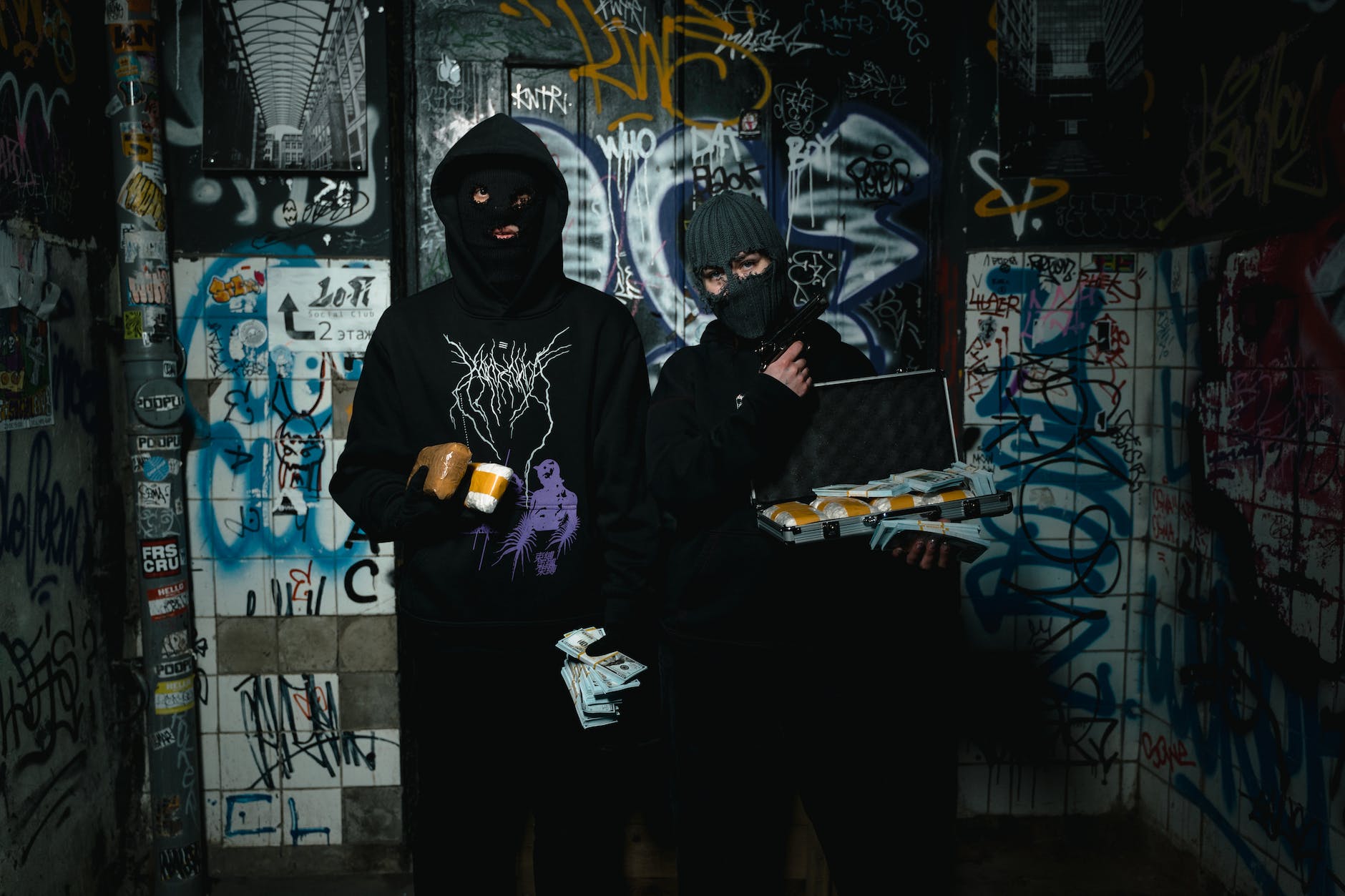 two people in black hoodie standing beside a wall with graffiti holding cash money